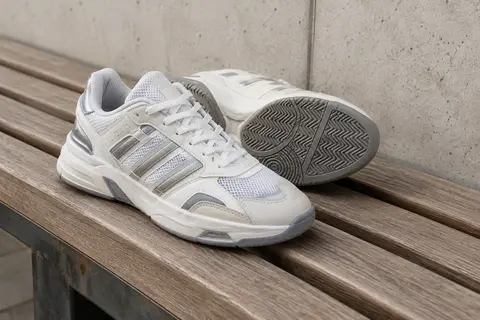 Paire de chaussures de basketball blanches et argentées, à lacets, posées sur un banc en bois.