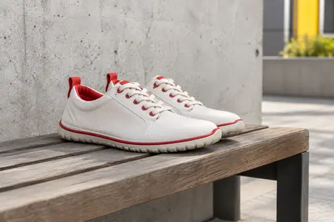Baskets barefoot Hobibear blanches et rouges en toile, posées sur un banc en bois devant un mur de béton clair.
