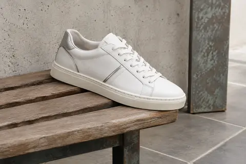 Sneaker basse Puma Carina L en cuir blanc et détails argentés, posée sur un banc en bois.