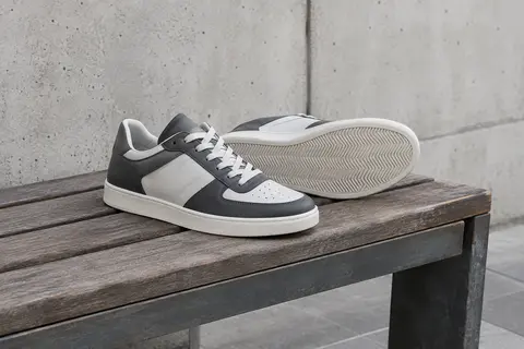 Paire de baskets basses gris foncé et blanches posées sur un banc en bois devant un mur de béton clair