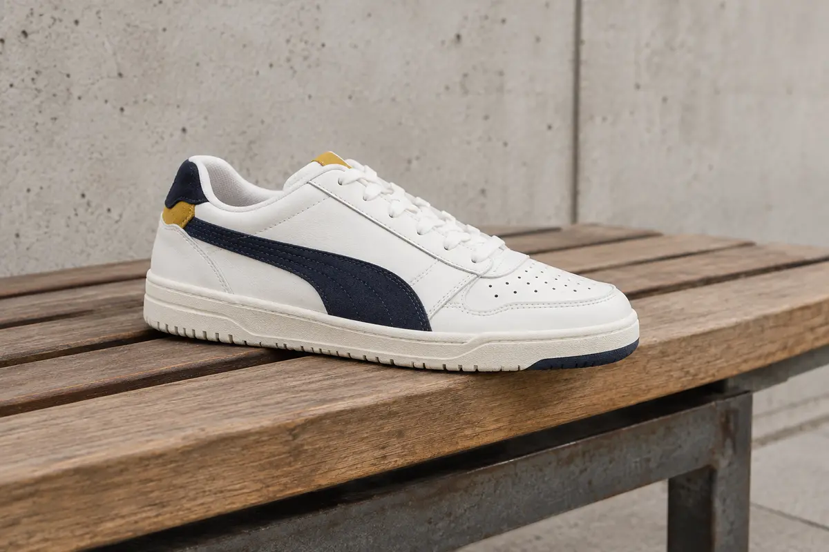 Basket Puma Shuffle basse blanche, avec touches Peacoat et Team Gold, posée sur un banc en bois.