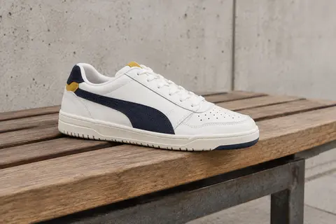 Basket Puma Shuffle basse blanche, avec touches Peacoat et Team Gold, posée sur un banc en bois.