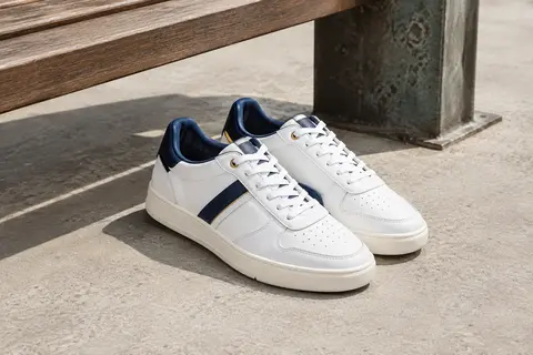Paire de sneakers basses en cuir blanc, bleu marine et doré, posées sur un sol de béton clair en ville.