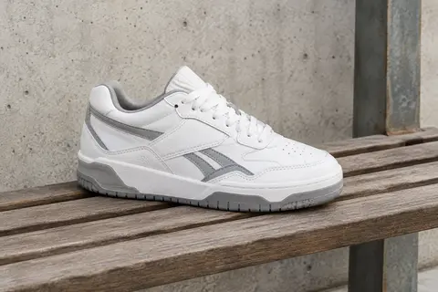Sneaker Reebok BB 1000 blanche et grise posée sur un banc en bois dans un décor urbain sobre.